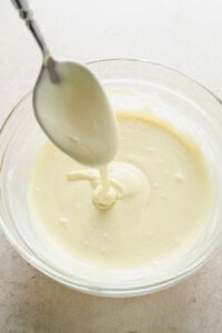 A spoon drizzling melted white chocolate into a clear glass bowl filled with more melted white chocolate.