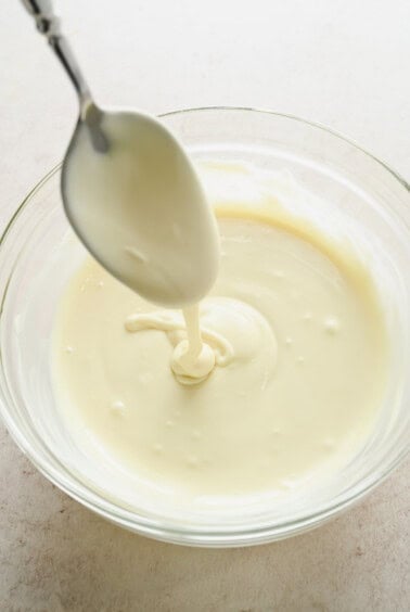 A spoon drizzling melted white chocolate into a clear glass bowl filled with more melted white chocolate.