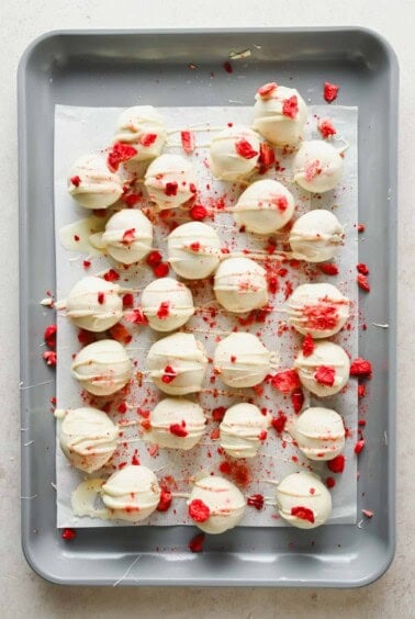 A baking sheet with white chocolate-coated round treats, drizzled with more white chocolate and sprinkled with crushed red pieces, arranged on parchment paper.