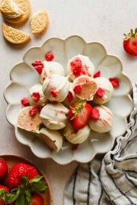 A plate of white chocolate-covered strawberry truffles is garnished with crumbled cookies and strawberry pieces; fresh strawberries and cookies are nearby.