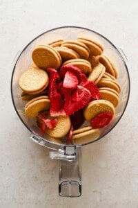 A food processor bowl filled with golden sandwich cookies and freeze-dried strawberry slices on a light surface.