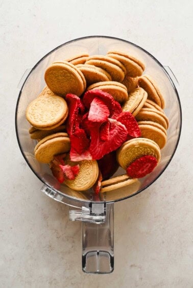 A food processor bowl filled with golden sandwich cookies and freeze-dried strawberry slices on a light surface.