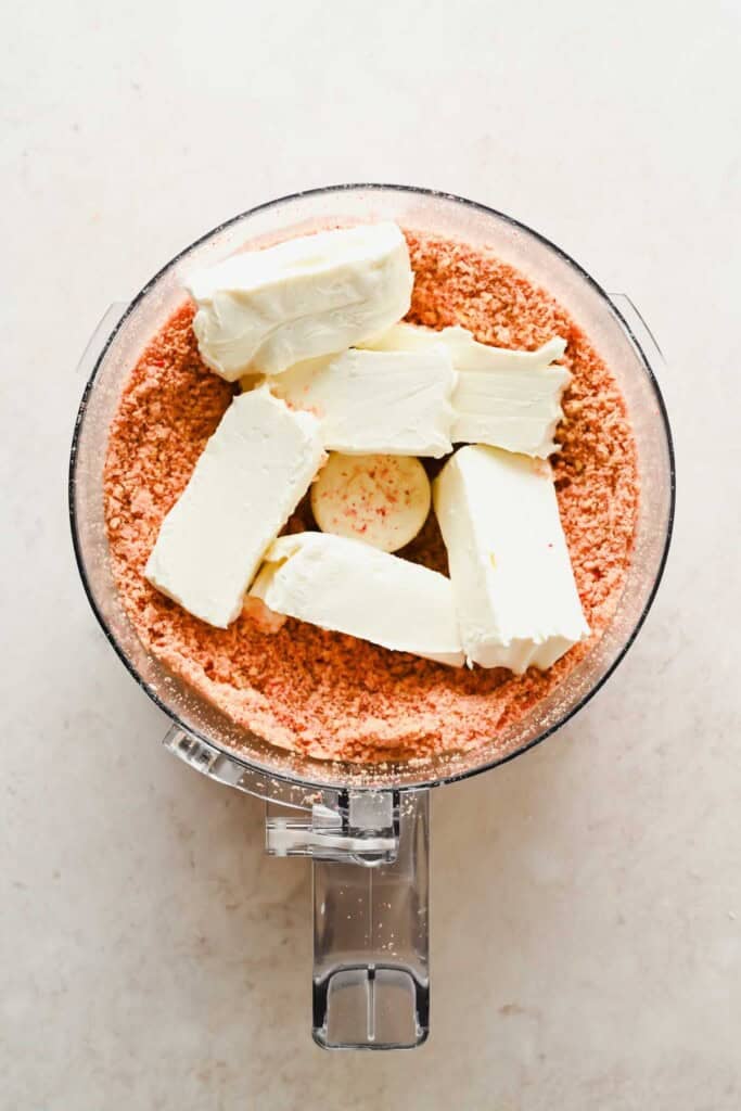 Food processor bowl with crushed pink cookies at the bottom and six blocks of cream cheese on top.