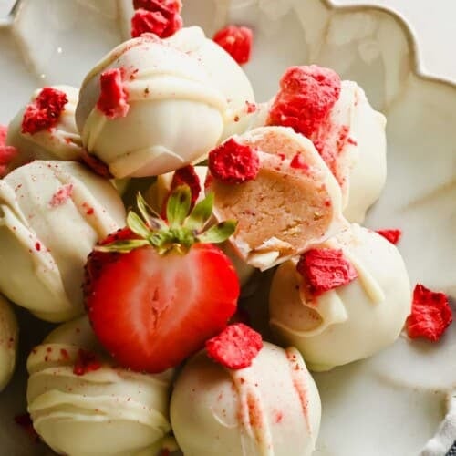 A plate of white chocolate-coated truffles, garnished with freeze-dried strawberry pieces and a halved fresh strawberry. One truffle is cut open to show the filling.