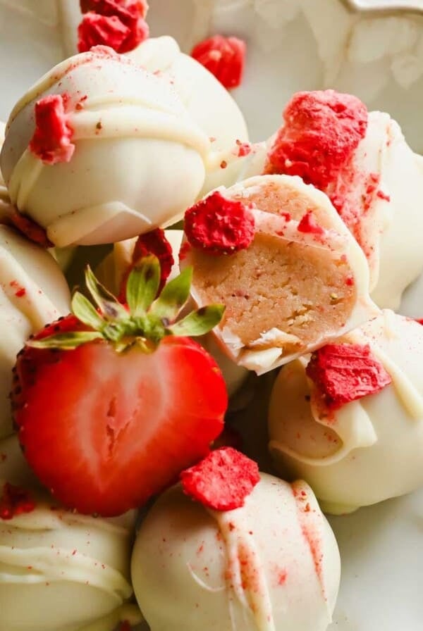A plate of white chocolate-coated truffles, garnished with freeze-dried strawberry pieces and a halved fresh strawberry. One truffle is cut open to show the filling.