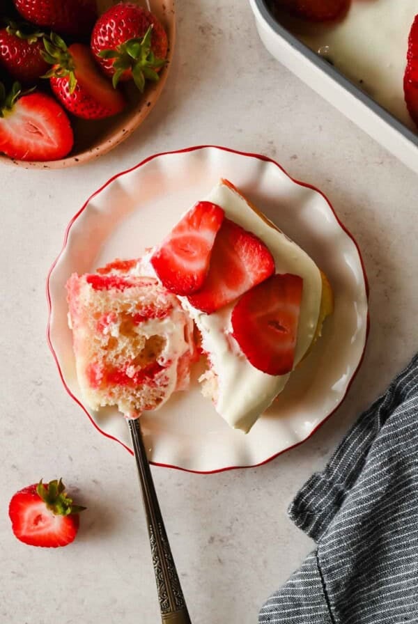 A slice of strawberry shortcake with cream frosting and fresh strawberry slices on a white plate, with a fork and whole strawberries nearby.