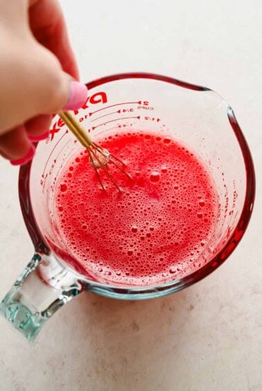 A hand uses a whisk to mix a red liquid in a glass measuring cup on a light-colored surface.