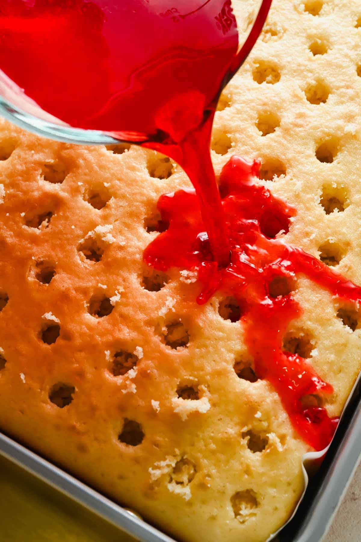 Pouring strawberry Jello mixture over a poked vanilla cake in a baking pan