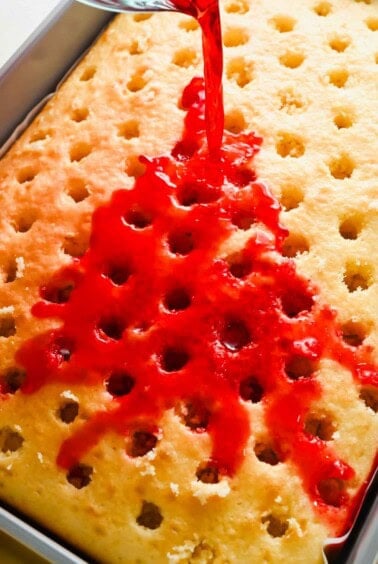 Red gelatin mixture being poured over a yellow sheet cake with holes poked in it, in a rectangular baking pan.