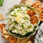 A bowl of chicken salad spread garnished with scallions and lemon, served with pretzel crisps and square crackers on the side.