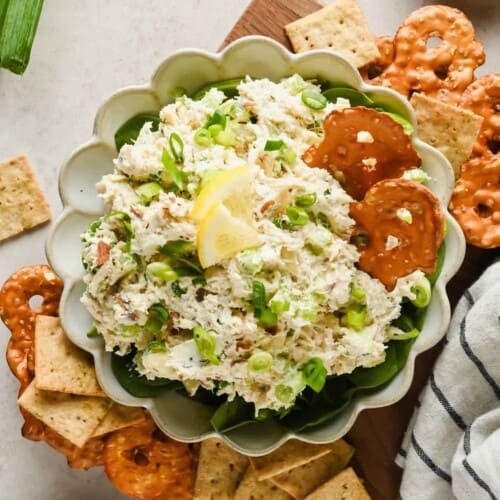 A bowl of chicken salad spread garnished with scallions and lemon, served with pretzel crisps and square crackers on the side.