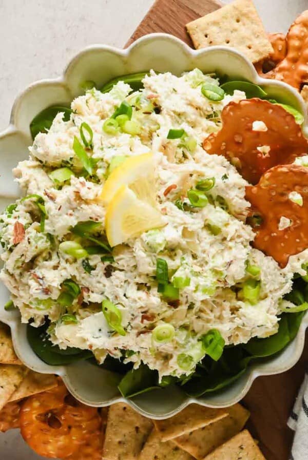 A bowl of chicken salad spread garnished with scallions and lemon, served with pretzel crisps and square crackers on the side.