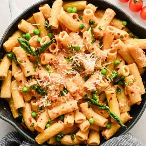 A skillet filled with rigatoni pasta in a creamy cottage cheese pasta sauce, topped with grated cheese, green peas, fresh basil, and red pepper flakes.