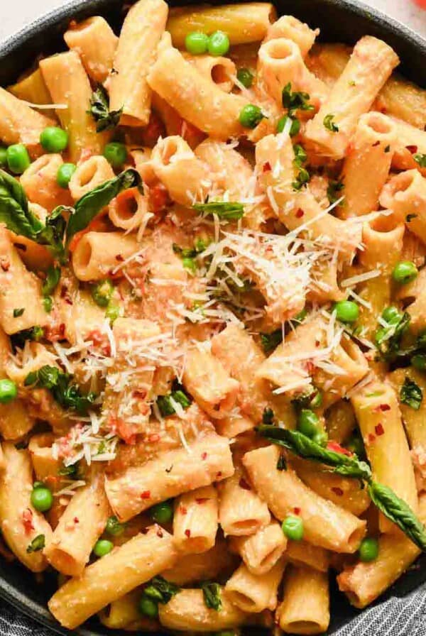 A skillet filled with rigatoni pasta in a creamy cottage cheese pasta sauce, topped with grated cheese, green peas, fresh basil, and red pepper flakes.