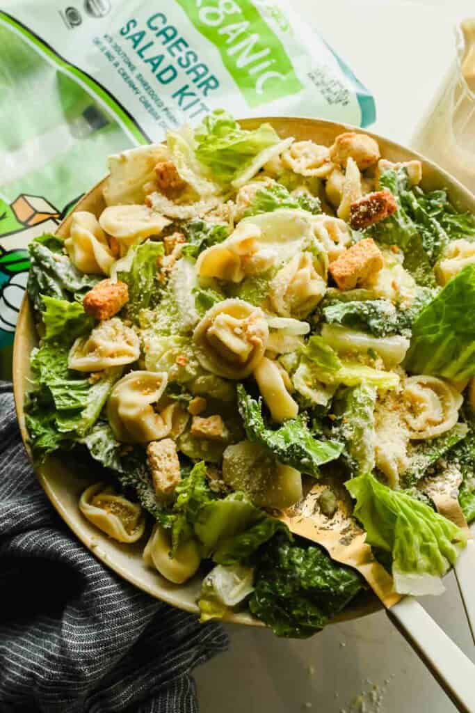 A delicious bowl of tortellini Caesar salad featuring tender tortellini, crisp croutons, and grated cheese on top, served next to a convenient salad kit package.