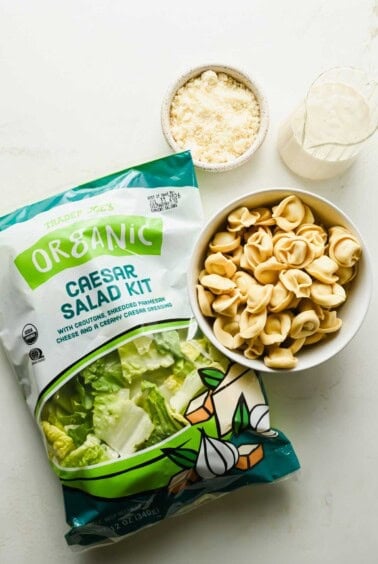 A bag of Trader Joe’s Organic Caesar Salad Kit, a bowl of tortellini caesar salad, grated parmesan in a small bowl, and a glass cup of creamy dressing on a white surface.