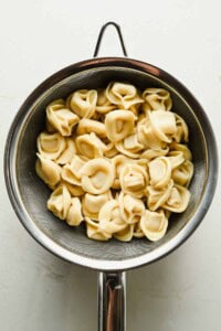 Cooked cheese tortellini, perfect for a refreshing tortellini caesar salad, draining in a metal colander over a white surface.