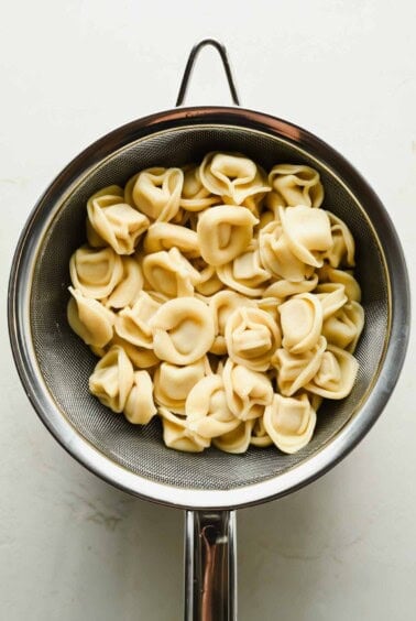 Cooked cheese tortellini, perfect for a refreshing tortellini caesar salad, draining in a metal colander over a white surface.