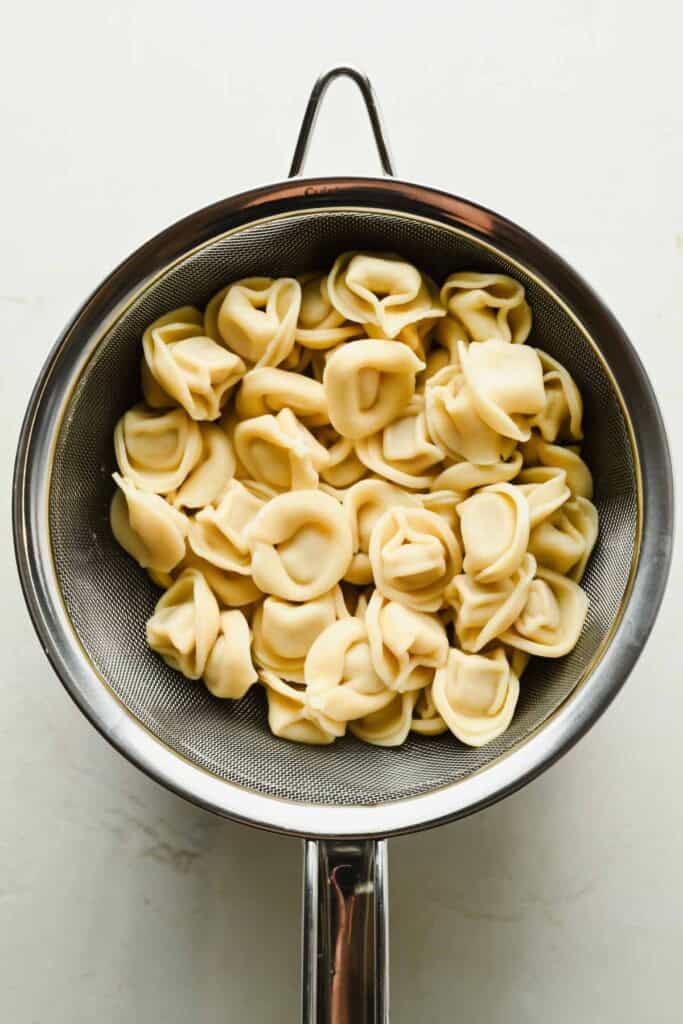 Cooked cheese tortellini, perfect for a refreshing tortellini caesar salad, draining in a metal colander over a white surface.