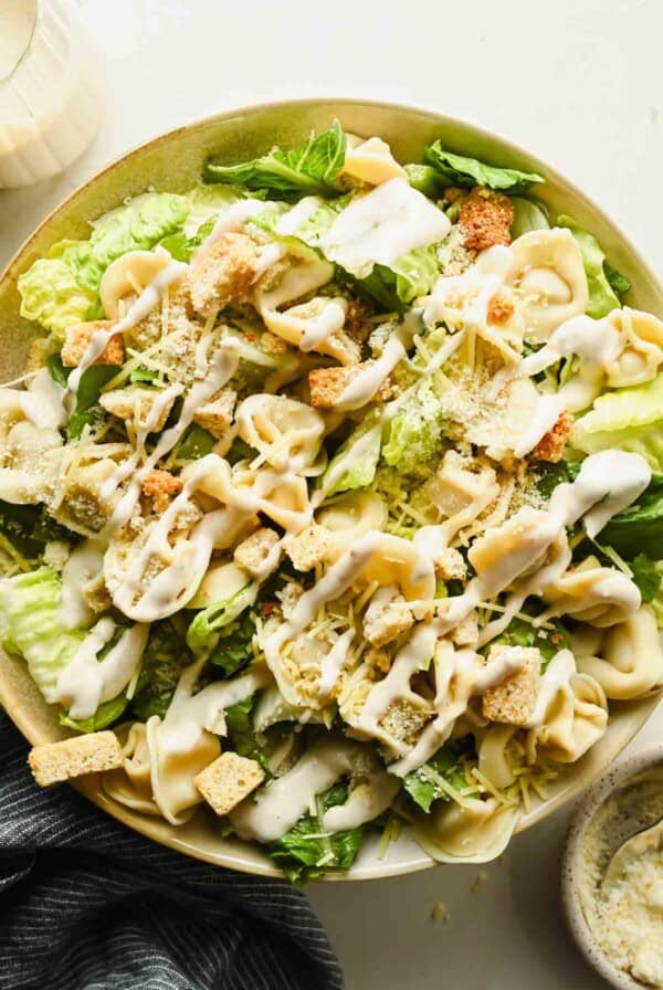 A bowl of tortellini Caesar salad featuring tender pasta, crunchy croutons, shredded cheese, and a generous drizzle of Caesar dressing on top.