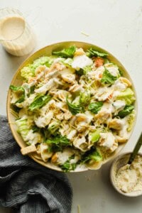 A bowl of tortellini Caesar salad featuring pasta, grilled chicken, croutons, fresh greens, and Caesar dressing, with a small pitcher of dressing and grated cheese nearby.