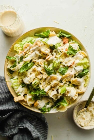 A bowl of tortellini Caesar salad featuring pasta, grilled chicken, croutons, fresh greens, and Caesar dressing, with a small pitcher of dressing and grated cheese nearby.