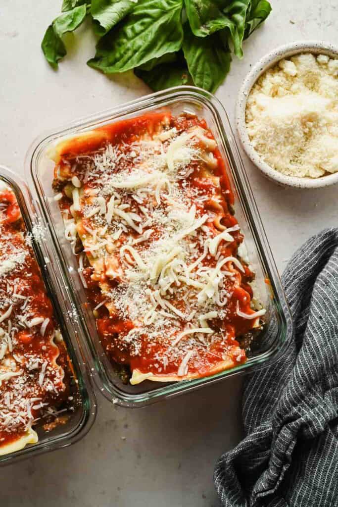 Two glass meal prep containers of microwave lasagna topped with marinara and melted cheese