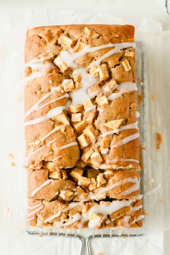 A loaf of apple bread topped with chopped apple pieces and drizzled with white icing sits on parchment paper and a cooling rack.