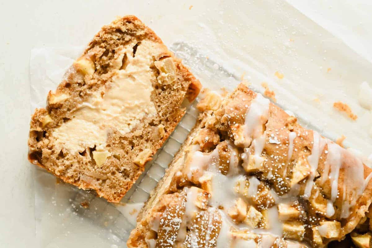 A sliced loaf of apple bread with visible apple pieces, cream cheese filling, and icing drizzled on top, sitting on parchment paper.