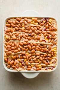 Accordion-folded tortillas topped with pinto beans, corn, and diced red onion in a square baking dish.
