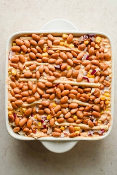 Accordion-folded tortillas topped with pinto beans, corn, and diced red onion in a square baking dish.