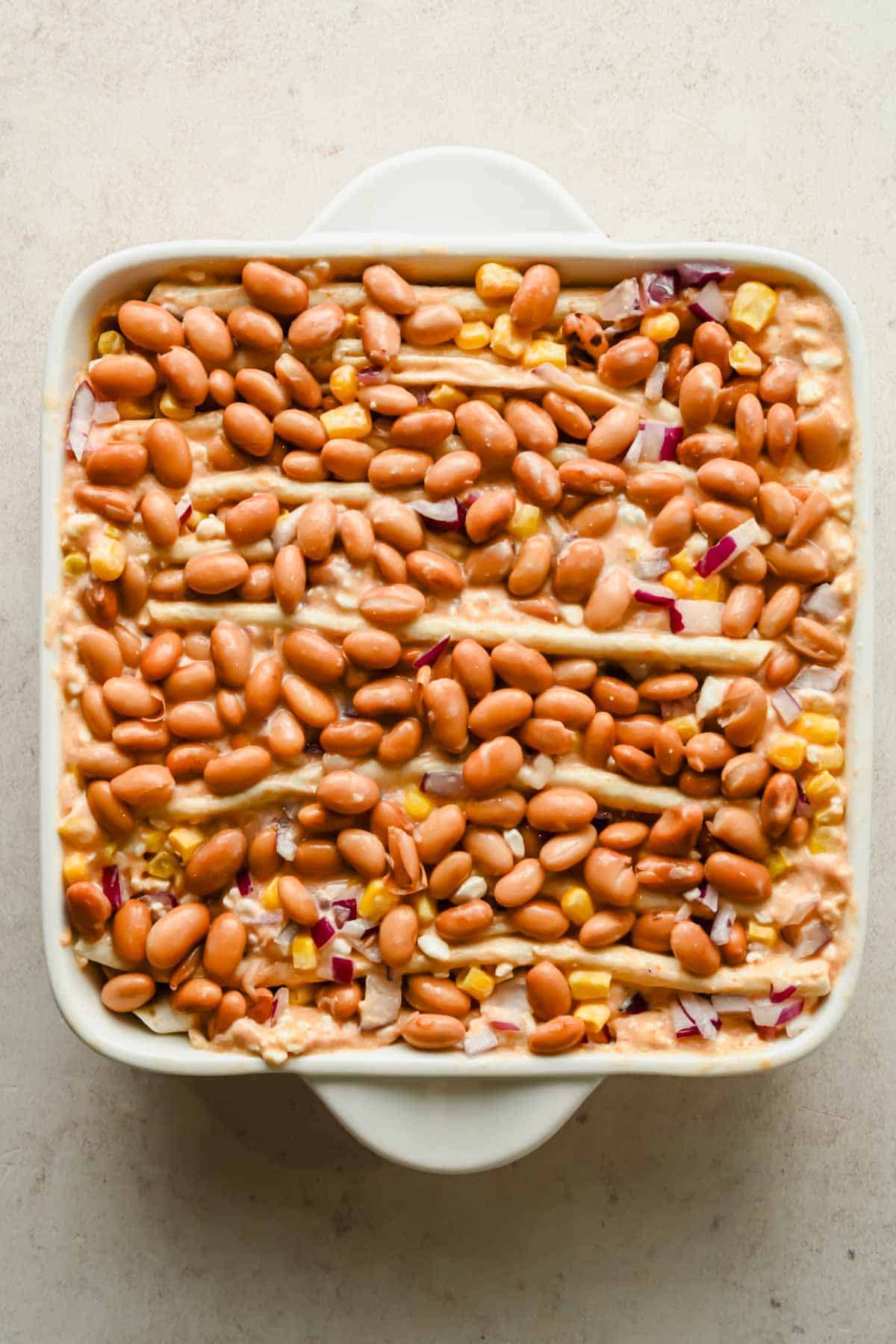 Accordion-folded tortillas topped with pinto beans, corn, and diced red onion in a square baking dish.