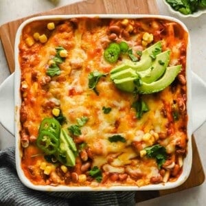 A baked casserole reminiscent of deconstructed enchiladas, topped with melted cheese, sliced avocado, jalapeño, corn, beans, and cilantro, served in a white square dish on a wooden board.