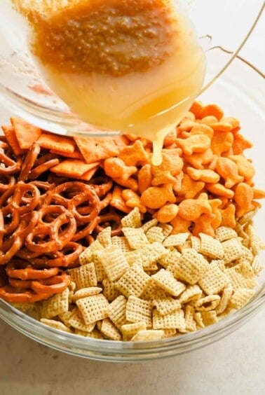 Seasoned melted butter being poured over Chex cereal, pretzels, and cheese crackers.