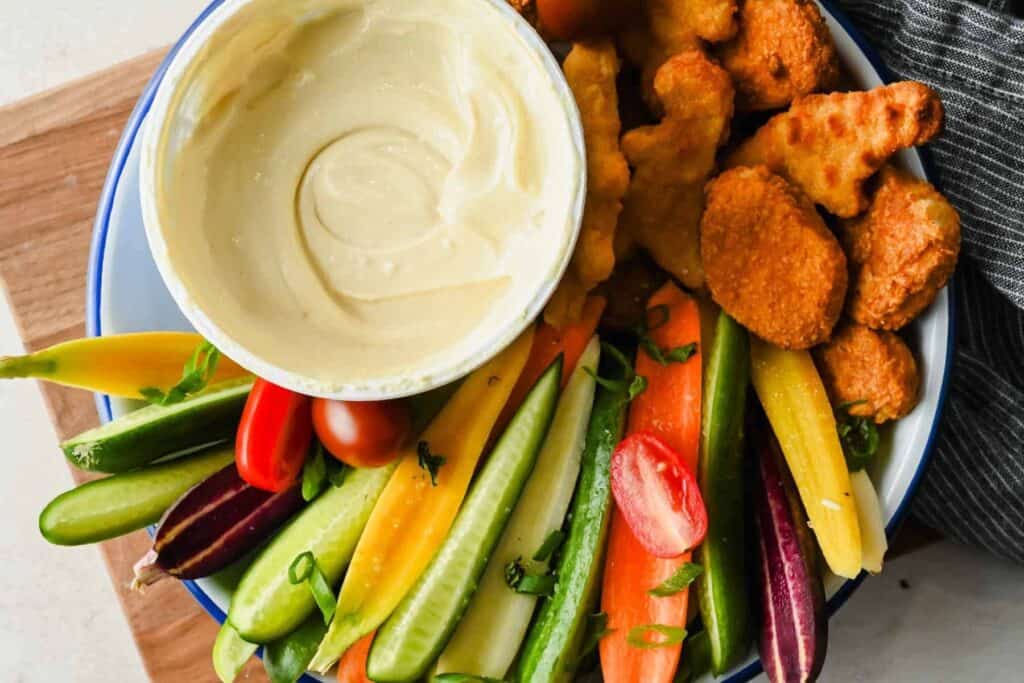 A bowl of cottage cheese honey mustard dip with chicken nuggets, carrots, cucumbers, and tomatoes.