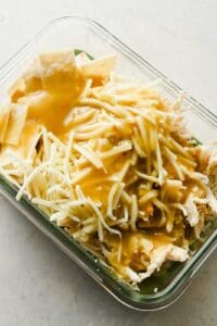 Torn tortilla, cooked chicken, green enchilada sauce, and Monterey Jack in a glass container.