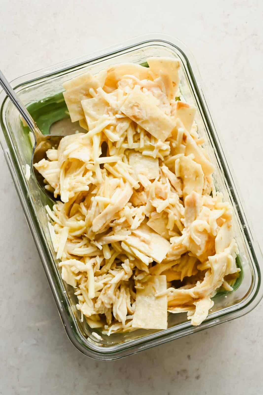 Torn tortilla, cooked chicken, green enchilada sauce, and Monterey Jack in a glass container.